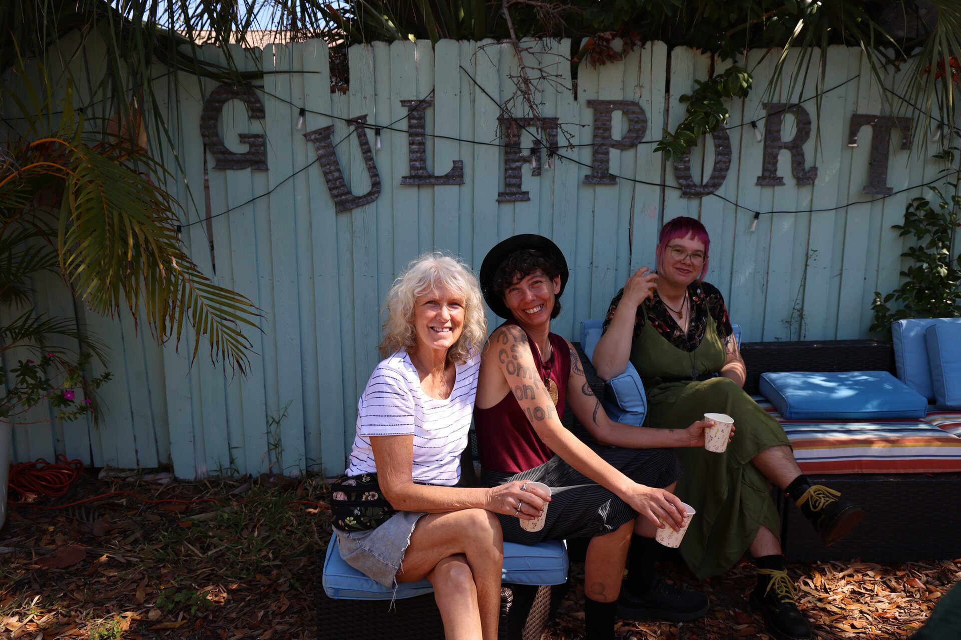 Three smiling women share drinks together in the garden in front of the Gulfport sign